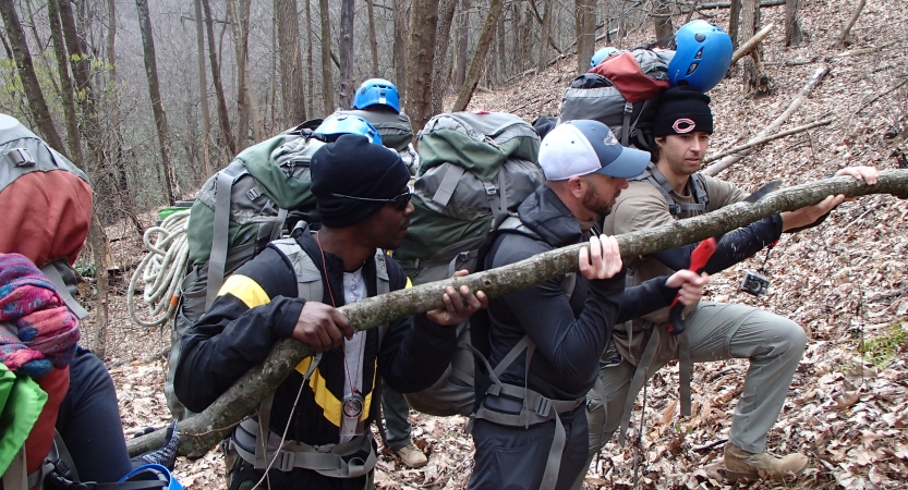 A group of people carry a large branch in a wooded environment. 